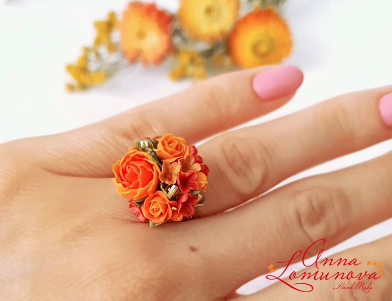 Peut inclure: Une bague orn&eacute;e d'un bouquet de fleurs en p&acirc;te polym&egrave;re orange et rouge. La bague est port&eacute;e au doigt.
