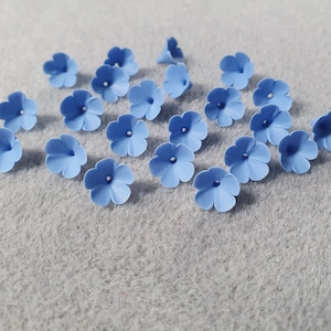 Puede incluir: Una colección de 18 adornos de flores de fieltro azul claro. Las flores son todas del mismo tamaño y forma, con cinco pétalos y un pequeño agujero en el centro.