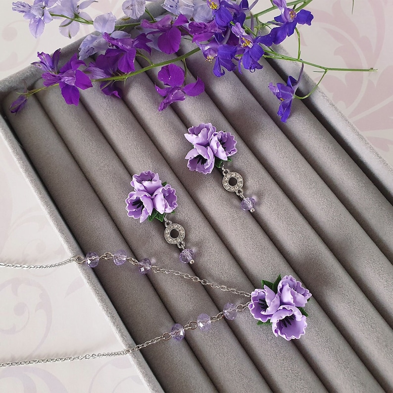 May include: A delicate silver necklace and earrings set with purple flower accents. The necklace features a chain with small purple beads and a larger flower pendant. The earrings are drop earrings with a smaller flower design.