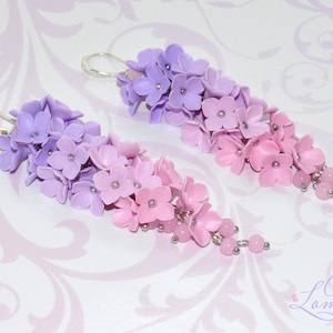 Peut inclure: Une paire de boucles d'oreilles pendantes avec un d&eacute;grad&eacute; de fleurs violettes et roses. Les fleurs sont en p&acirc;te polym&egrave;re et sont enfil&eacute;es sur un fil d'argent. Les boucles d'oreilles sont termin&eacute;es par de petites perles roses.