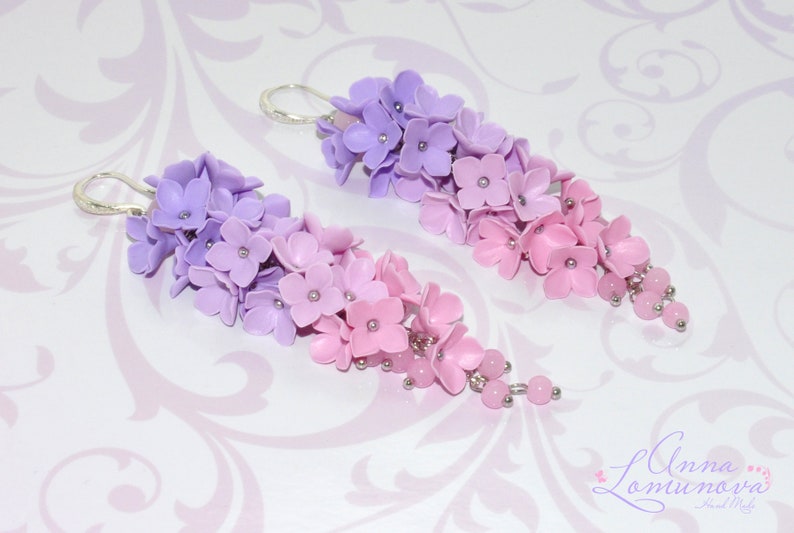Peut inclure: Une paire de boucles d'oreilles pendantes avec un d&eacute;grad&eacute; de fleurs violettes et roses. Les fleurs sont en p&acirc;te polym&egrave;re et sont enfil&eacute;es sur un fil d'argent. Les boucles d'oreilles sont termin&eacute;es par de petites perles roses.
