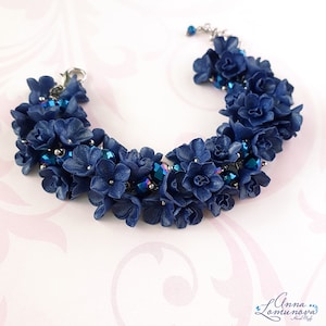 以下が含まれることがあります： ネイビーブルーの花のアクセントと虹色の青いクリスタルビーズが特徴の手作りのブレスレット。ブレスレットはシルバーカラーの留め金とチェーンで作られています。花の要素は一緒にクラスター化され、完全で質感のある外観を作り出しています。