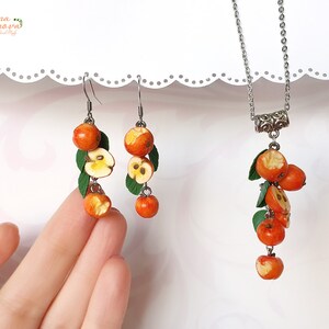 May include: A set of earrings and a necklace featuring a cluster of small, red, and yellow polymer clay apples with green leaves. The jewelry is made with silver-toned metal.