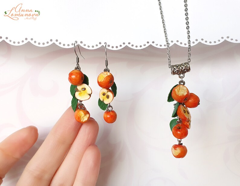 May include: A set of earrings and a necklace featuring a cluster of small, red, and yellow polymer clay apples with green leaves. The jewelry is made with silver-toned metal.