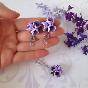 May include: A set of lavender flower earrings and a matching necklace. The earrings feature a silver ring with clear rhinestones and a dangling crystal. The necklace has a silver chain and a lavender flower pendant.