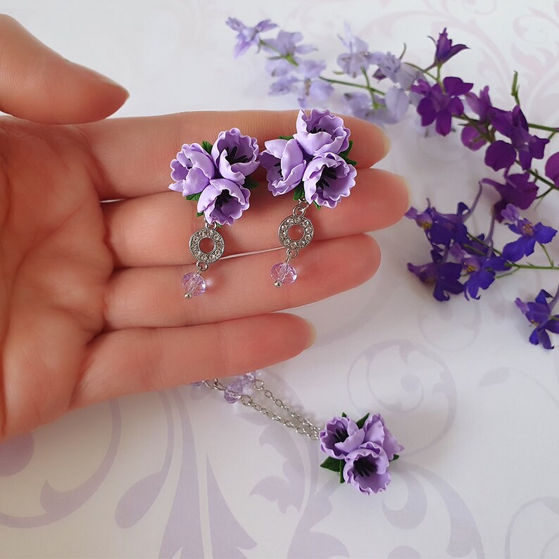 May include: A set of lavender flower earrings and a matching necklace. The earrings feature a silver ring with clear rhinestones and a dangling crystal. The necklace has a silver chain and a lavender flower pendant.
