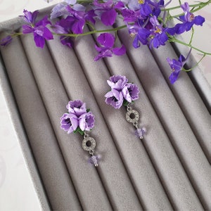 May include: A pair of purple flower earrings with a silver ring and a purple crystal dangle. The earrings are displayed on a grey velvet background.