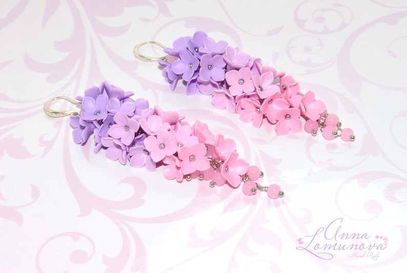 Peut inclure: Une paire de boucles d'oreilles pendantes avec un d&eacute;grad&eacute; de fleurs violettes et roses. Les fleurs sont en p&acirc;te polym&egrave;re et sont enfil&eacute;es sur un fil d'argent. Les boucles d'oreilles sont finies avec de petites perles argent&eacute;es.