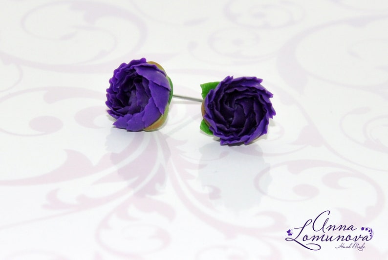 May include: A pair of purple flower stud earrings with green leaves. The earrings are made of polymer clay and are handmade.