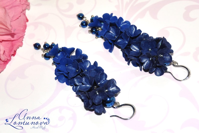 May include: A pair of handmade, dark blue floral earrings. Each earring features a cascade of small, textured flower petals and a cluster of dark blue beads. The earrings have silver-colored, curved ear wires.
