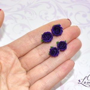 May include: Four small purple fabric roses with green leaves. The roses are arranged in a row on a hand.