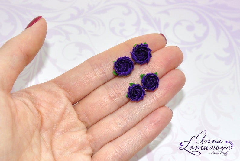 May include: Four small purple fabric roses with green leaves. The roses are arranged in a row on a hand.