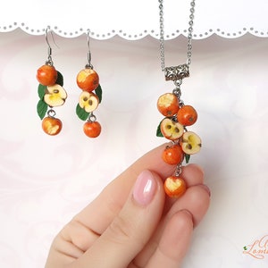 May include: A set of earrings and a necklace featuring a cluster of red and yellow apple charms. The charms are made to look like cut apples with visible seeds. The jewelry is made with silver-toned metal.
