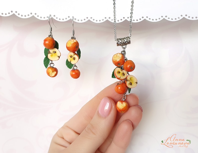 May include: A set of earrings and a necklace featuring a cluster of red and yellow apple charms. The charms are made to look like cut apples with visible seeds. The jewelry is made with silver-toned metal.