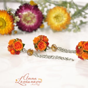 Peut inclure: Une paire de boucles d'oreilles en forme de fleurs orange et rouge avec des cha&icirc;nes argent&eacute;es. Les boucles d'oreilles sont en p&acirc;te polym&egrave;re et ont la forme de petits bouquets de fleurs.