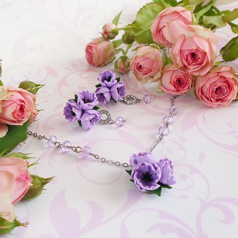 May include: A delicate silver necklace and earrings set with purple flower accents. The necklace features a cluster of three flowers and the earrings each have three flowers.
