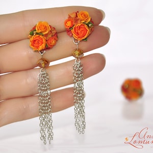 Peut inclure: Une paire de boucles d'oreilles en forme de fleurs orange et rouge avec des cha&icirc;nes argent&eacute;es. Les boucles d'oreilles sont tenues dans une main.