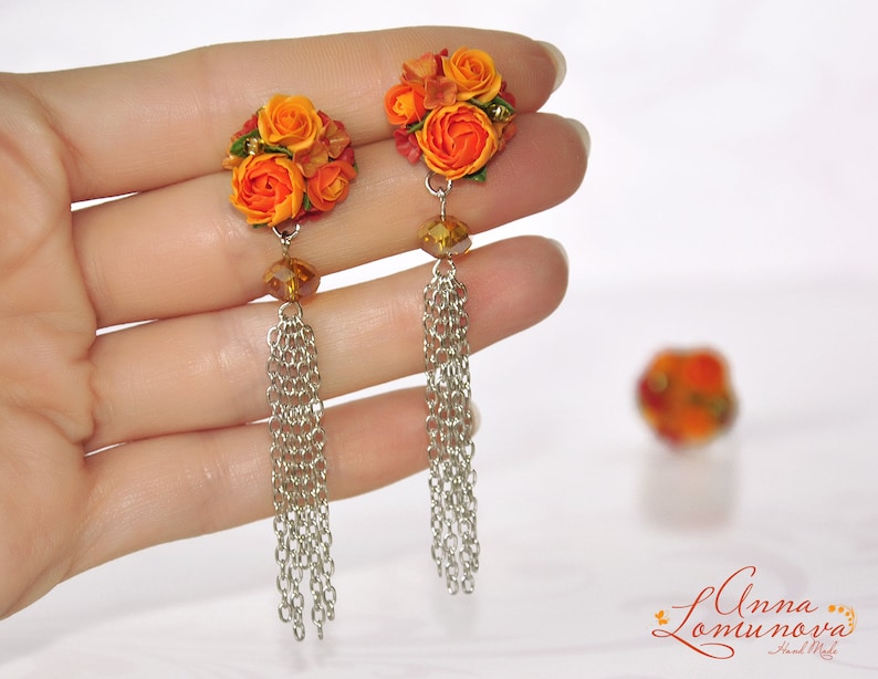 Peut inclure: Une paire de boucles d'oreilles en forme de fleurs orange et rouge avec des cha&icirc;nes argent&eacute;es. Les boucles d'oreilles sont tenues dans une main.