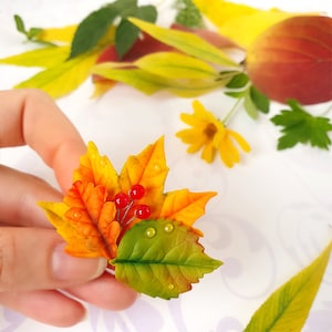 Op de afbeelding: Een handgemaakte broche van polymer clay met een cluster van herfstbladeren in tinten oranje, geel en groen. De bladeren zijn versierd met kleine rode bessen en hebben een realistisch, getextureerd uiterlijk.
