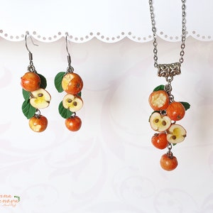 May include: A set of earrings and a necklace featuring a cluster of red and orange apple charms with green leaves. The charms are made of polymer clay and are shaped like whole apples and apple halves.