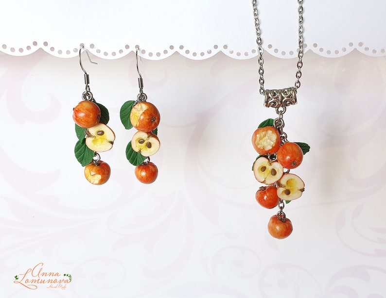 May include: A set of earrings and a necklace featuring a cluster of red and orange apple charms with green leaves. The charms are made of polymer clay and are shaped like whole apples and apple halves.