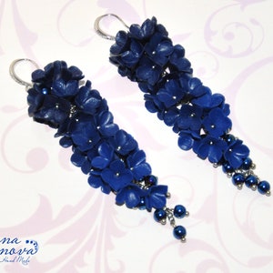 May include: A pair of long, dangling earrings featuring clusters of dark blue flower-shaped beads. The earrings have silver-colored hooks and small, dark blue beads at the bottom. The earrings are handmade.