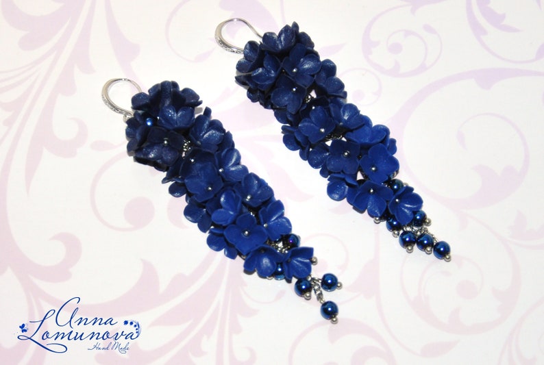 May include: A pair of long, dangling earrings featuring clusters of dark blue flower-shaped beads. The earrings have silver-colored hooks and small, dark blue beads at the bottom. The earrings are handmade.
