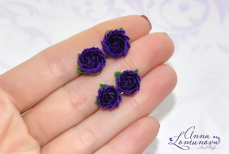 May include: Four small purple polymer clay flowers with green leaves. The flowers are arranged in a row on a hand.