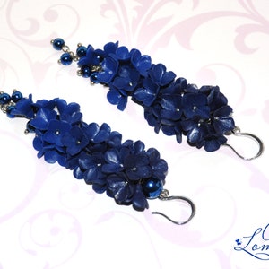 May include: A pair of long, dangling earrings featuring clusters of dark blue, flower-shaped beads. Each earring has a silver-colored hook and is accented with small, dark blue beads. The earrings are handmade.