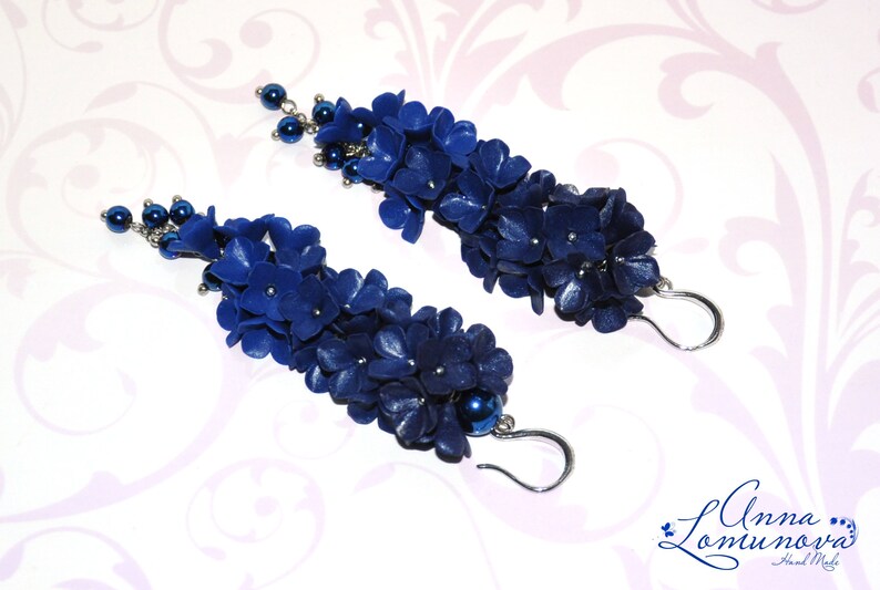 May include: A pair of long, dangling earrings featuring clusters of dark blue, flower-shaped beads. Each earring has a silver-colored hook and is accented with small, dark blue beads. The earrings are handmade.
