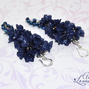 May include: A pair of long, dangling earrings with navy blue flower-shaped charms and silver hooks. The charms are clustered together and have a sparkly, textured finish. The earrings are accented with small, dark blue beads.