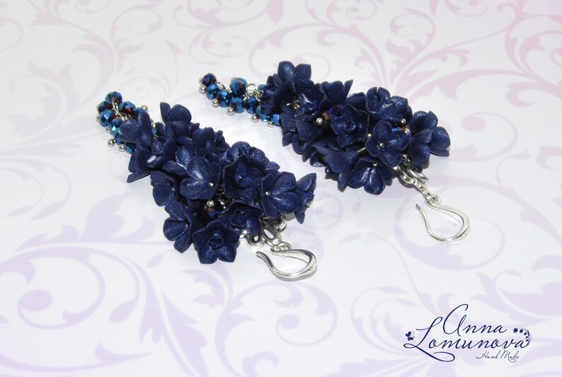 May include: A pair of long, dangling earrings with navy blue flower-shaped charms and silver hooks. The charms are clustered together and have a sparkly, textured finish. The earrings are accented with small, dark blue beads.