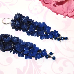 May include: A pair of long, dangling earrings featuring clusters of deep blue flowers and small, dark blue beads. The earrings have silver-colored hooks and are set against a patterned background with pink flowers.