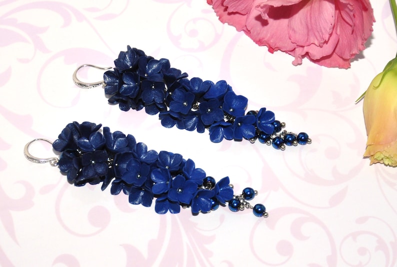 May include: A pair of long, dangling earrings featuring clusters of deep blue flowers and small, dark blue beads. The earrings have silver-colored hooks and are set against a patterned background with pink flowers.