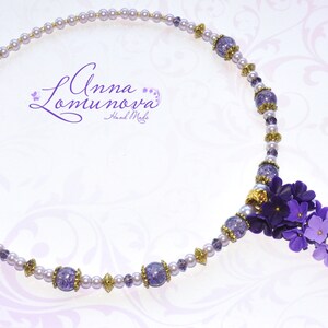 Peut inclure: Un collier d&eacute;licat de couleur violet et blanc avec un bouquet de fleurs violettes qui pend en bas. Le collier est compos&eacute; de petites perles nacr&eacute;es et d'accents dor&eacute;s. Le texte "Anna Lomunova Hand Made" est visible sur le collier.