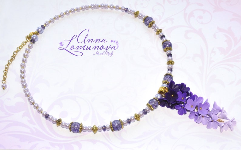 Peut inclure: Un collier d&eacute;licat de couleur violet et blanc avec un bouquet de fleurs violettes qui pend en bas. Le collier est compos&eacute; de petites perles nacr&eacute;es et d'accents dor&eacute;s. Le texte "Anna Lomunova Hand Made" est visible sur le collier.