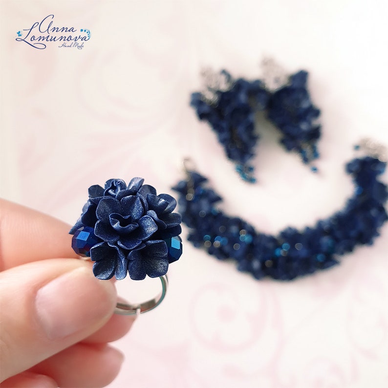 以下が含まれることがあります： 手作りのジュエリーセット。濃い青色の花の指輪、お揃いのイヤリング、ブレスレットが含まれています。指輪は、大きな花のモチーフとファセットブルーのアクセントが特徴です。イヤリングとブレスレットにも花のモチーフがあります。