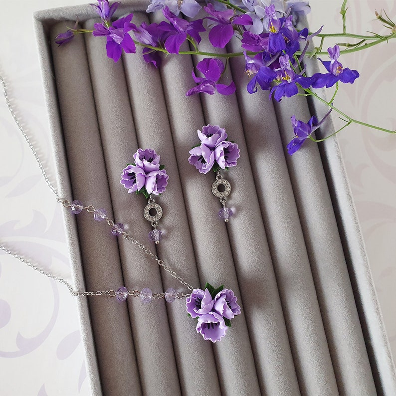 May include: A delicate necklace and earrings set featuring clusters of purple flowers with silver accents. The jewellery is displayed in a grey velvet box.