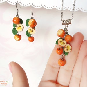 May include: A silver necklace and matching earrings feature a cluster of red and yellow polymer clay apples with green leaves. The necklace has a silver chain and a decorative silver clasp.