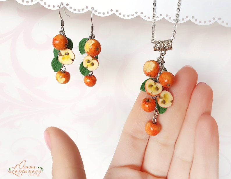 May include: A silver necklace and matching earrings feature a cluster of red and yellow polymer clay apples with green leaves. The necklace has a silver chain and a decorative silver clasp.