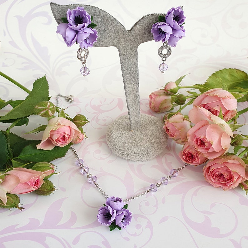 May include: A delicate lilac flower necklace and earrings set. The necklace features a cluster of three lilac flowers with crystal accents. The earrings feature a single lilac flower with a crystal dangle.