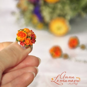 Peut inclure: Une bague orn&eacute;e d'un petit bouquet de fleurs en p&acirc;te polym&egrave;re orange et rouge. Les fleurs sont regroup&eacute;es en grappe et entour&eacute;es de feuilles vertes. La bague est port&eacute;e &agrave; un doigt.