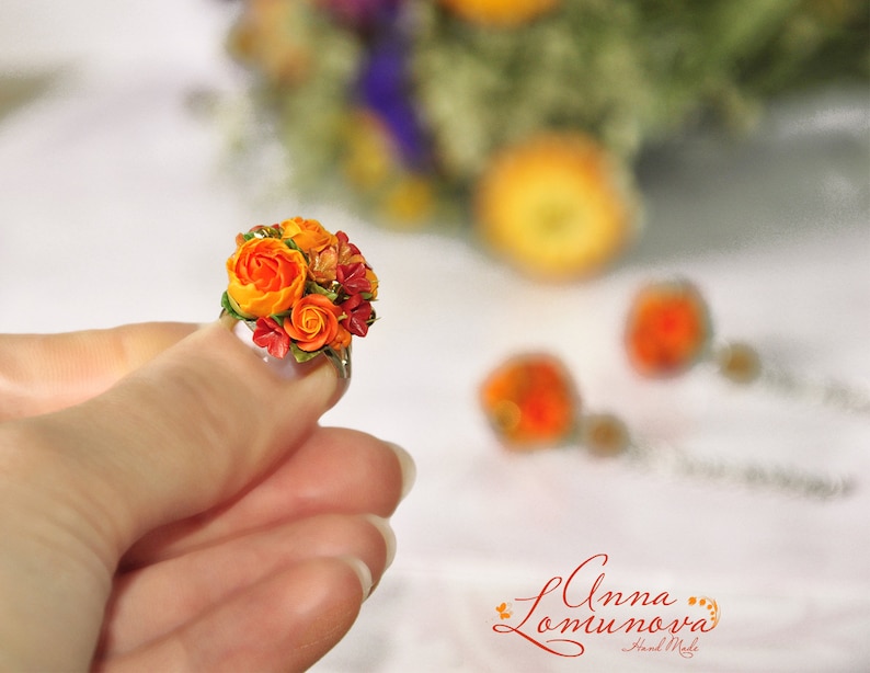 Peut inclure: Une bague orn&eacute;e d'un petit bouquet de fleurs en p&acirc;te polym&egrave;re orange et rouge. Les fleurs sont regroup&eacute;es en grappe et entour&eacute;es de feuilles vertes. La bague est port&eacute;e &agrave; un doigt.