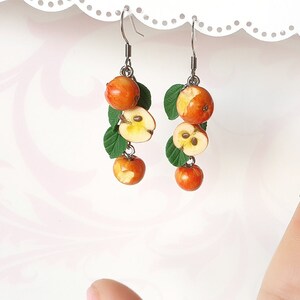 May include: Handmade earrings featuring miniature orange apples, some sliced to reveal the seeds, and green leaves. The earrings hang from silver-colored hooks. A unique, nature-inspired jewelry design.