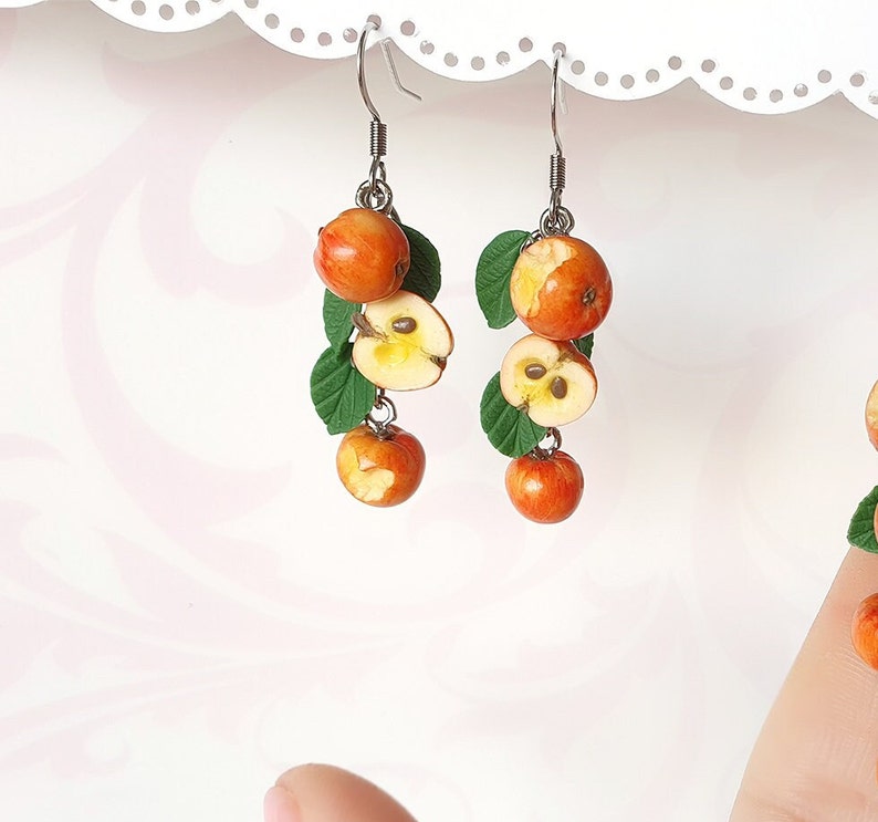 May include: Handmade earrings featuring miniature orange apples, some sliced to reveal the seeds, and green leaves. The earrings hang from silver-colored hooks. A unique, nature-inspired jewelry design.