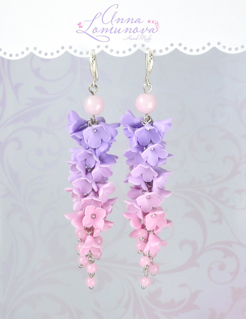 Peut inclure: Une paire de boucles d'oreilles pendantes avec un d&eacute;grad&eacute; de fleurs en p&acirc;te polym&egrave;re violettes et roses. Les boucles d'oreilles sont faites avec des crochets et des perles argent&eacute;s.