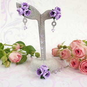 May include: A delicate purple flower necklace and earrings set with crystal accents. The necklace features a cluster of three purple flowers and the earrings each have a cluster of three purple flowers.