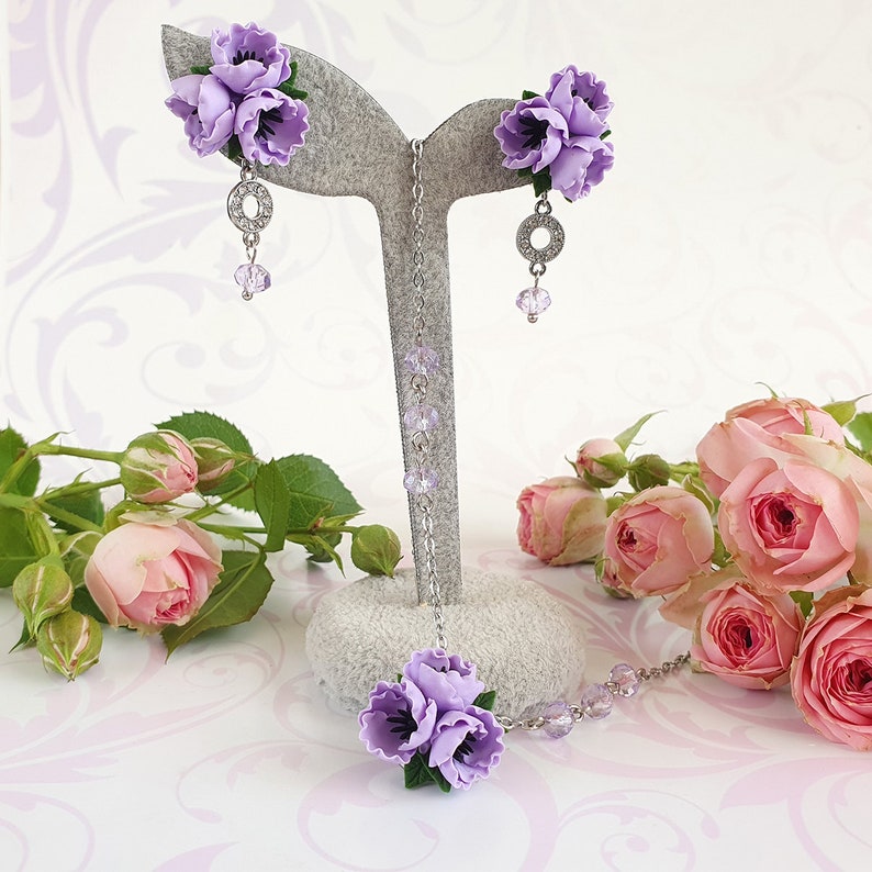 May include: A delicate purple flower necklace and earrings set with crystal accents. The necklace features a cluster of three purple flowers and the earrings each have a cluster of three purple flowers.