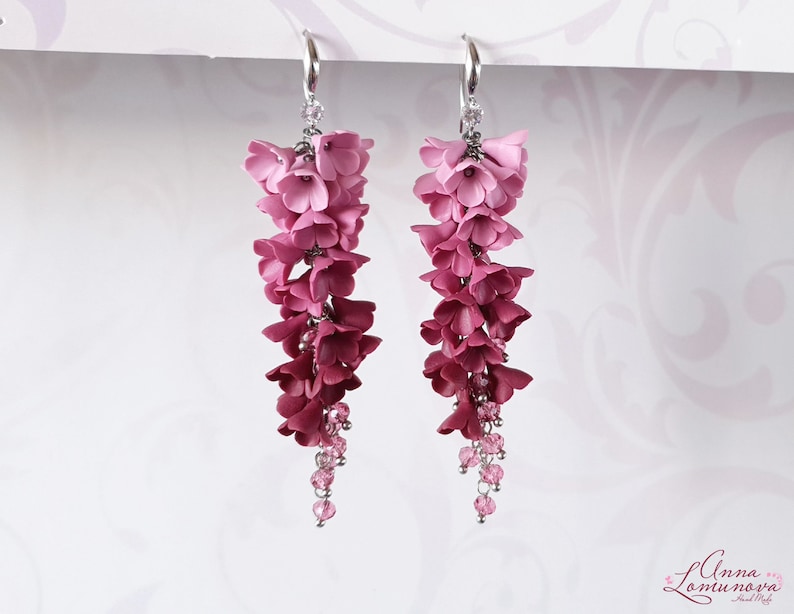 May include: A pair of dangle earrings with a gradient of pink and red polymer clay flowers. The flowers are arranged in a cascading design and are accented with small pink crystal beads.