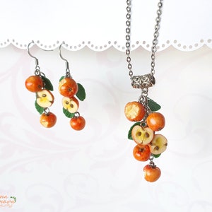 May include: A jewelry set featuring a necklace and earrings with a fruit theme. The necklace has a silver chain with a pendant of miniature orange apples, some sliced to reveal the interior. The earrings mirror the design, with green leaves.